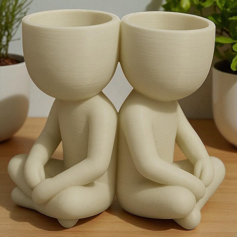 May include: Two off-white ceramic planters shaped like seated figures with their legs crossed. Each figure has a large, open head that serves as a planting pot. The figures are positioned back-to-back, creating a unique decorative piece.