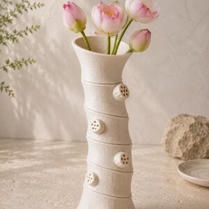 May include: An off-white ceramic vase with a textured finish, holding pink and white lotus flowers. The vase has a distinctive design with circular embellishments. The vase is a decorative home accent, and is approximately 30 cm tall.