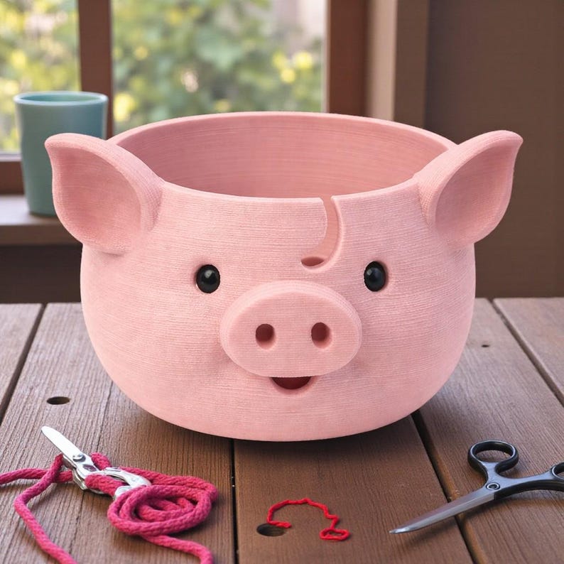 Puede incluir: Una maceta rosa con forma de cerdo con ojos negros y un hocico sonriente. La maceta tiene orejas grandes y una superficie texturizada. Unas tijeras y hilo rosa est&aacute;n sobre la superficie de madera frente a la maceta.