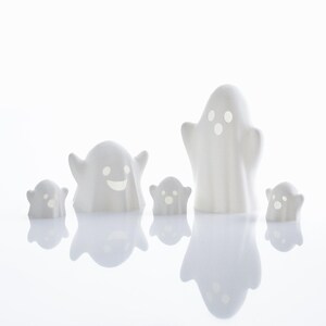 May include: Five white ceramic ghost figurines with different facial expressions. The ghosts are arranged in a row on a white surface.