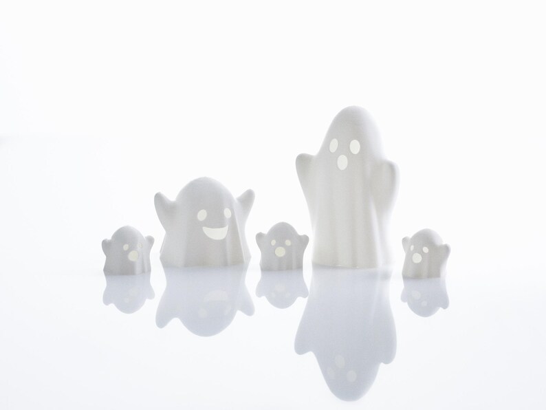 May include: Five white ceramic ghost figurines with different facial expressions. The ghosts are arranged in a row on a white surface.