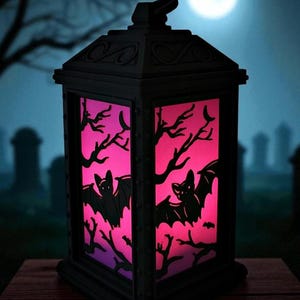 May include: A black Halloween lantern with a pink and purple illuminated interior. The lantern features bat and tree branch cutouts. The background shows a graveyard scene with a full moon.