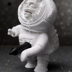 Pu&ograve; includere: Figurina bianca stampata in 3D di un astronauta con un casco danneggiato che rivela una faccia mostruosa. Un oggetto cilindrico nero &egrave; inserito nel petto dell'astronauta. Su sfondo scuro.
