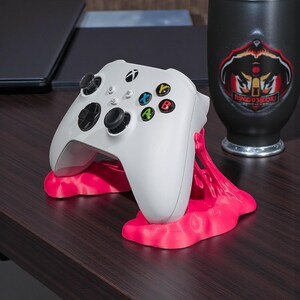 May include: A white Xbox controller rests on a bright pink stand. The controller has black joysticks and colored buttons. A black mug with a logo is visible in the background. The items are on a dark wood surface.