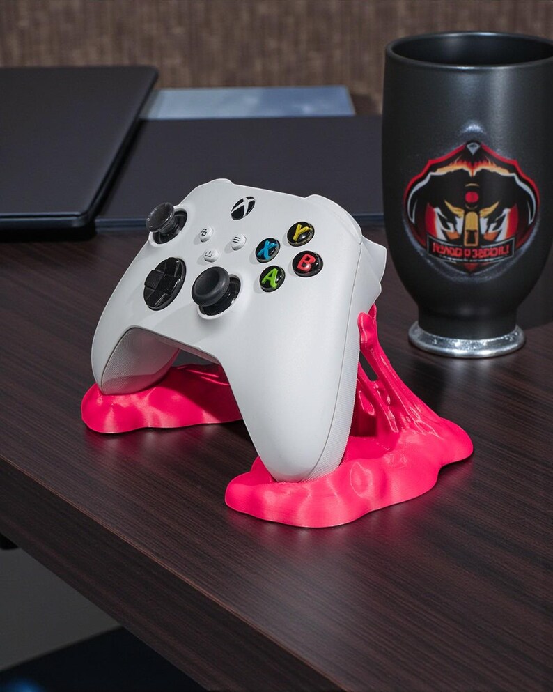 May include: A white Xbox controller rests on a bright pink stand. The controller has black joysticks and colored buttons. A black mug with a logo is visible in the background. The items are on a dark wood surface.