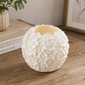 May include: A spherical, off-white candle holder with a floral design. The surface is covered in small, three-dimensional flowers. The interior is a lighter cream color. The candle holder sits on a wooden surface.