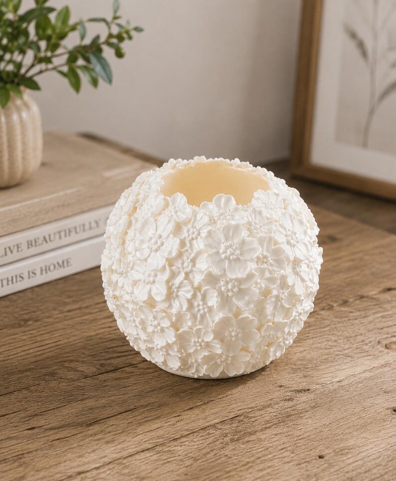 May include: A spherical, off-white candle holder with a floral design. The surface is covered in small, three-dimensional flowers. The interior is a lighter cream color. The candle holder sits on a wooden surface.