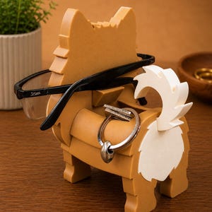 May include: A tan and white wooden dog-shaped organizer holding black-framed glasses and a silver keyring. The organizer has a slot for glasses and a ring holder. A small plant in a white pot is in the background.