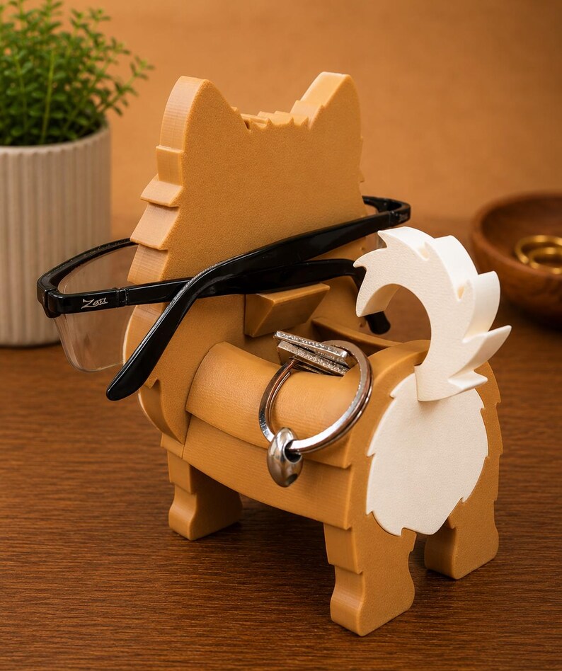 May include: A tan and white wooden dog-shaped organizer holding black-framed glasses and a silver keyring. The organizer has a slot for glasses and a ring holder. A small plant in a white pot is in the background.