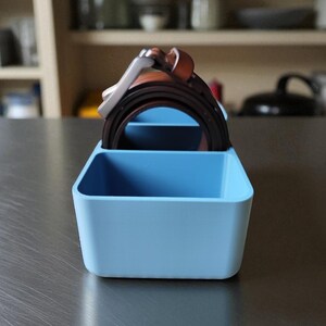 Puede incluir: Un organizador rectangular azul claro con dos compartimentos, que contiene varios cinturones marrones y negros. El organizador es de pl&aacute;stico y se encuentra sobre una superficie met&aacute;lica. Los cinturones tienen hebillas plateadas y est&aacute;n bien ordenados.