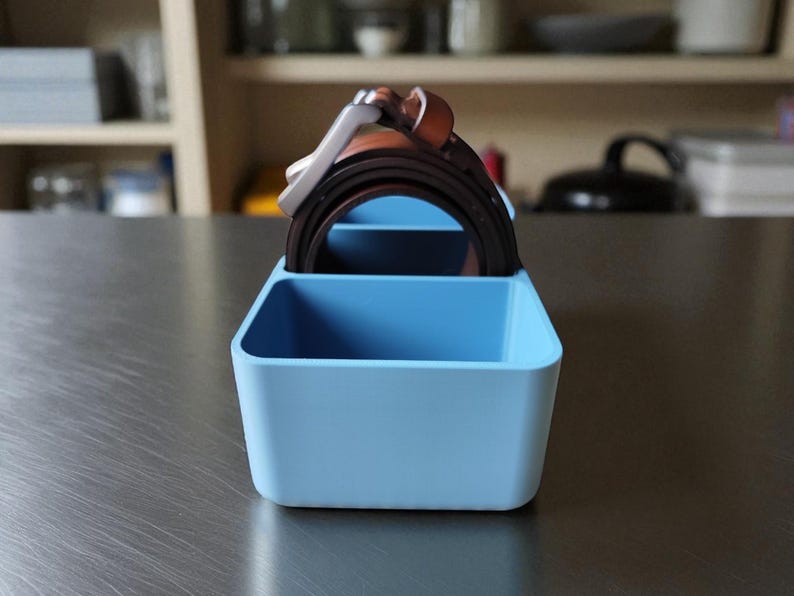 Puede incluir: Un organizador rectangular azul claro con dos compartimentos, que contiene varios cinturones marrones y negros. El organizador es de pl&aacute;stico y se encuentra sobre una superficie met&aacute;lica. Los cinturones tienen hebillas plateadas y est&aacute;n bien ordenados.