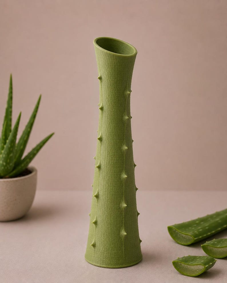May include: A green, textured vase with a unique design, resembling an aloe plant. The vase has a conical shape with a slanted opening and is adorned with small, pointed protrusions. The image also shows aloe vera leaves.