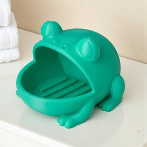 May include: A teal frog-shaped soap dish. The dish has a wide open mouth and a ridged interior. The frog has large, round eyes and small feet. The soap dish is made of a solid, smooth material and is designed to hold a bar of soap.