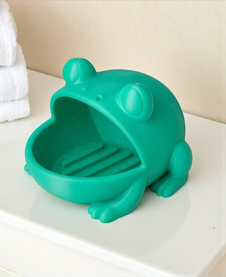May include: A teal frog-shaped soap dish. The dish has a wide open mouth and a ridged interior. The frog has large, round eyes and small feet. The soap dish is made of a solid, smooth material and is designed to hold a bar of soap.