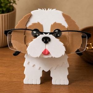 May include: A whimsical eyeglasses holder shaped like a Shih Tzu dog. The holder is white and brown with black eyes and a pink tongue, designed to hold a pair of eyeglasses. The dog is wearing black-framed glasses.