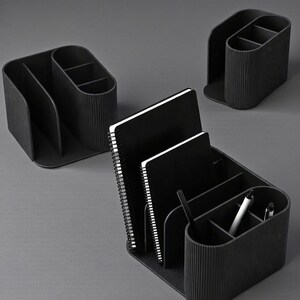 May include: Black desk organizers with multiple compartments and a ribbed texture. The organizers hold notebooks and pens. The design is modern and minimalist, suitable for office or home use.