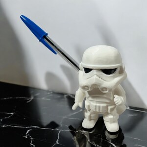 May include: A white Stormtrooper figurine with black eye coverings, holding a blue and clear pen. The figurine has detailed armor and stands on a black and white marbled surface. The pen is angled upwards.