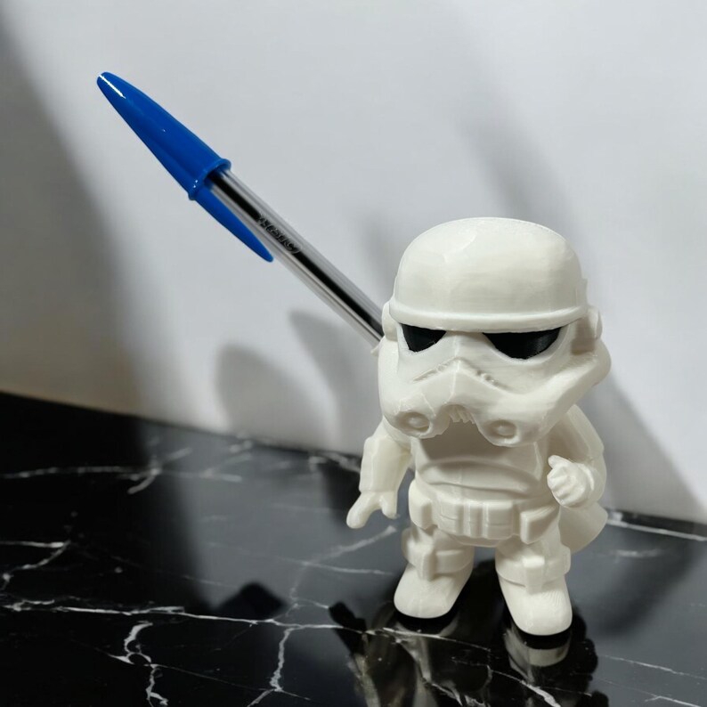 May include: A white Stormtrooper figurine with black eye coverings, holding a blue and clear pen. The figurine has detailed armor and stands on a black and white marbled surface. The pen is angled upwards.