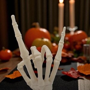 May include: A white, skeletal hand sculpture in the "rock on" gesture. The hand is the focal point, set against a blurred background of pumpkins, candles, and fall foliage. The scene evokes a Halloween or autumn theme.