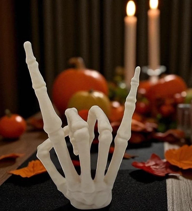 May include: A white, skeletal hand sculpture in the "rock on" gesture. The hand is the focal point, set against a blurred background of pumpkins, candles, and fall foliage. The scene evokes a Halloween or autumn theme.
