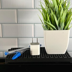 May include: A white wall charger with two prongs sits on a black grid surface next to two blue pens. A potted green plant with a white pot is in the background.