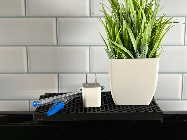 May include: A white wall charger with two prongs sits on a black grid surface next to two blue pens. A potted green plant with a white pot is in the background.