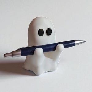May include: A white ghost-shaped pen holder with black eyes, holding a dark blue pen with a silver tip. The pen holder is a whimsical desk accessory.