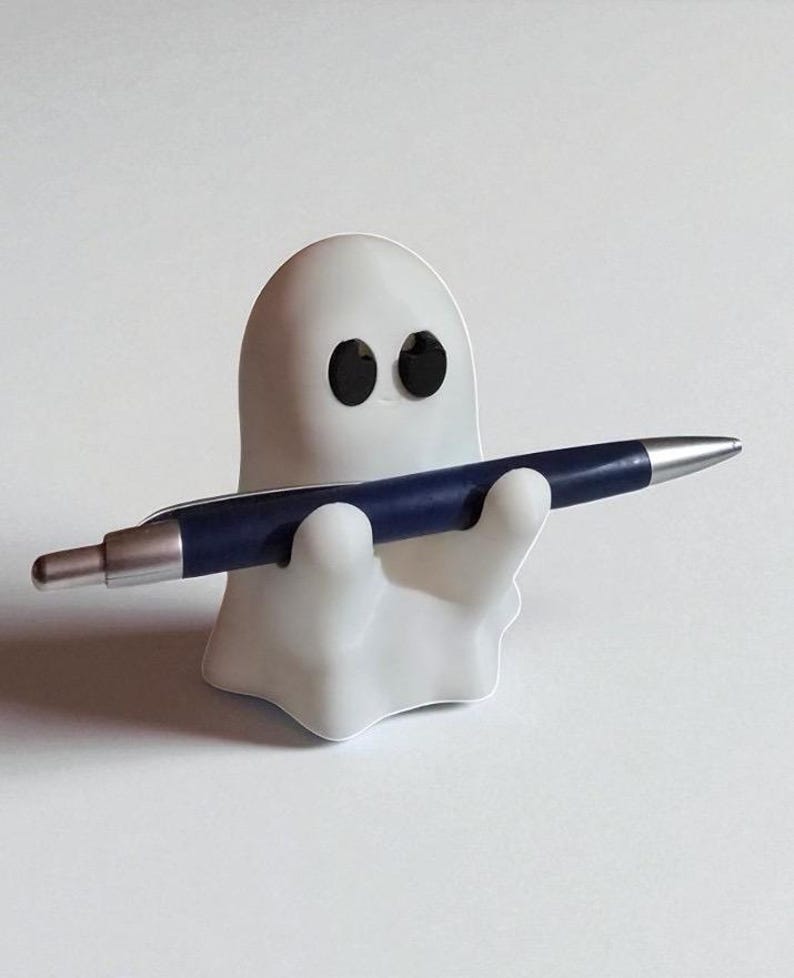 May include: A white ghost-shaped pen holder with black eyes, holding a dark blue pen with a silver tip. The pen holder is a whimsical desk accessory.