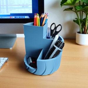 Op de afbeelding: Een lichtblauwe bureau-organizer met meerdere compartimenten. De organizer bevat kleurpotloden, een schaar, een pen en een stift. De organizer staat op een houten bureau, met een computermonitor en een plant op de achtergrond.