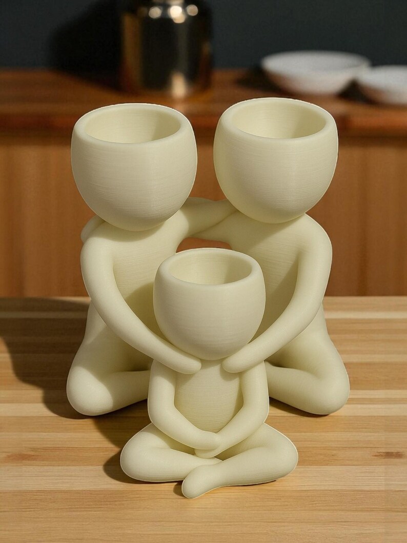 Puede incluir: Figurita familiar de color crema. Dos figuras abrazan a una figura m&aacute;s peque&ntilde;a, todas con macetas con forma de cabeza abierta. Las figuras est&aacute;n posadas sobre una superficie de madera. El objeto es decorativo.