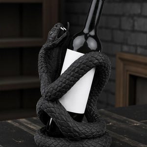 Puede incluir: Un soporte negro para botellas de vino con una serpiente texturizada detallada enrollada alrededor de una botella de vino oscura con una etiqueta blanca en blanco. La cabeza de la serpiente est&aacute; levantada, con la boca abierta. El soporte est&aacute; sobre una superficie de madera oscura.