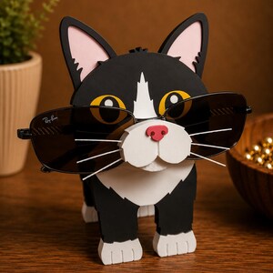 May include: A black and white cat-shaped sunglasses holder with pink ears and a red nose. The cat is wearing a pair of black sunglasses. The sunglasses have the Ray-Ban logo on the lens.