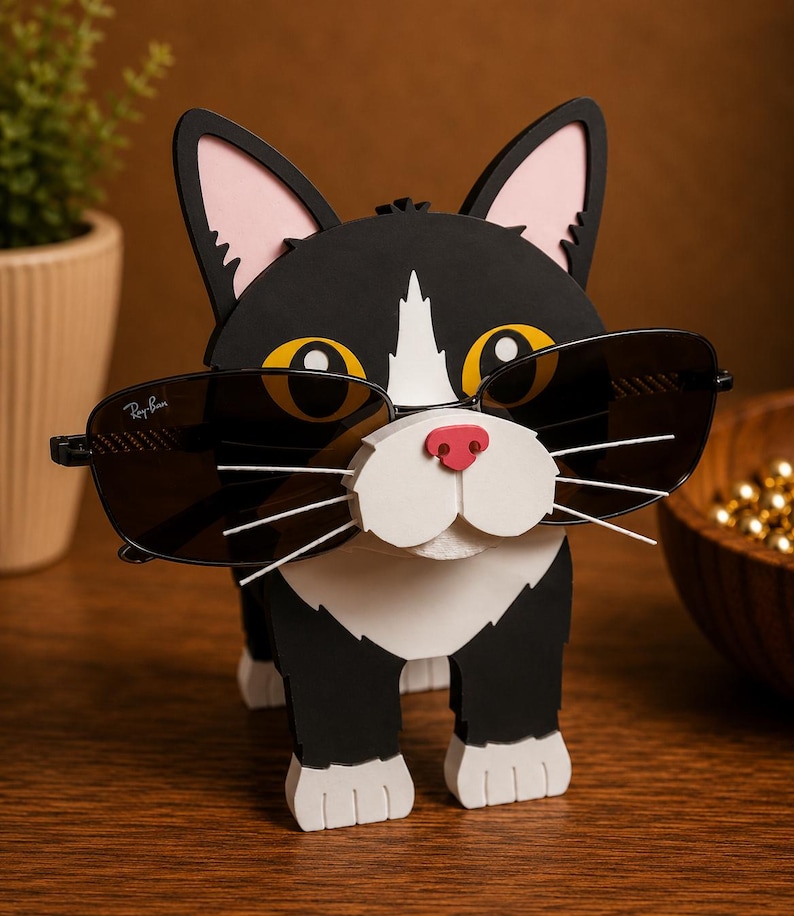 May include: A black and white cat-shaped sunglasses holder with pink ears and a red nose. The cat is wearing a pair of black sunglasses. The sunglasses have the Ray-Ban logo on the lens.