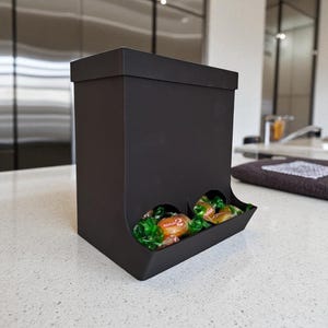 May include: A black candy dispenser with two dispensing slots. The dispenser is filled with wrapped candies. The dispenser is rectangular with a lid and sits on a white countertop. The candies are green and orange.