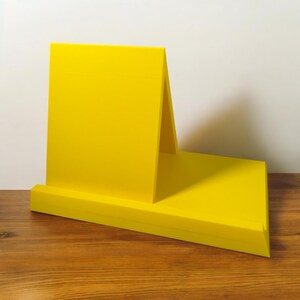 May include: A bright yellow wooden desk organizer with a slanted back and a flat base. The organizer is designed to hold items like notebooks or tablets. The design is modern and minimalist, with clean lines and a solid color.