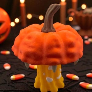 May include: A Halloween-themed decorative pumpkin with a unique design. The orange pumpkin has a textured surface, a brown stem, and two fangs. It sits atop a yellow base with white accents. Candy corn is scattered around the base.