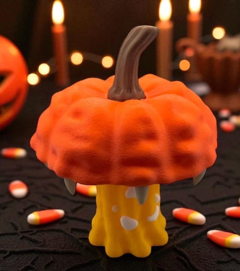 May include: A Halloween-themed decorative pumpkin with a unique design. The orange pumpkin has a textured surface, a brown stem, and two fangs. It sits atop a yellow base with white accents. Candy corn is scattered around the base.