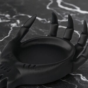 May include: A black decorative hand-shaped dish with a textured surface. The dish has a shallow bowl and is designed to hold small items. The fingers are long and pointed, giving it a gothic aesthetic.