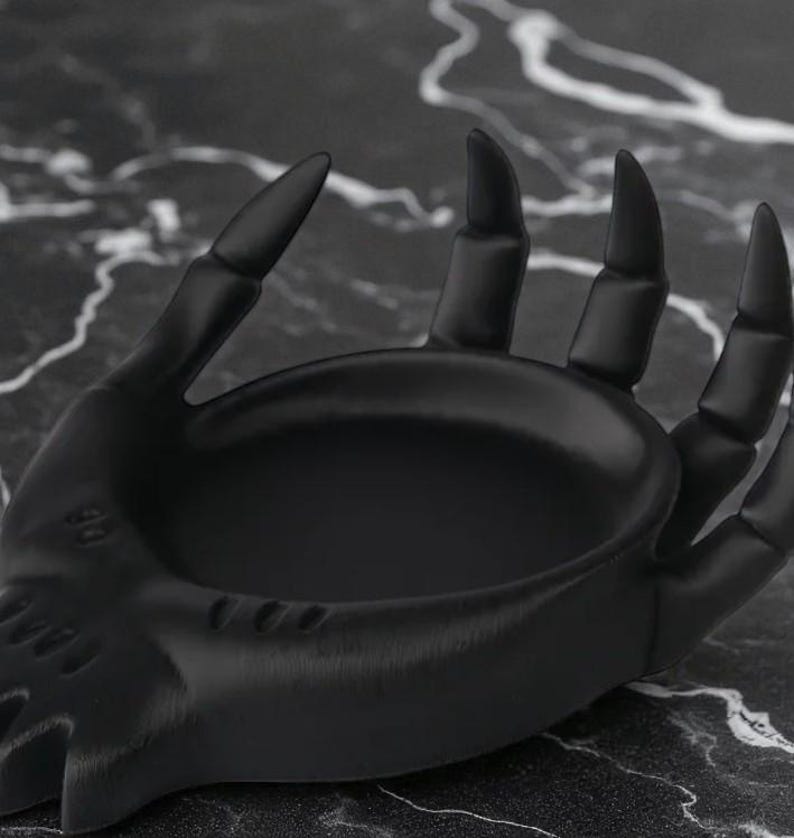 May include: A black decorative hand-shaped dish with a textured surface. The dish has a shallow bowl and is designed to hold small items. The fingers are long and pointed, giving it a gothic aesthetic.
