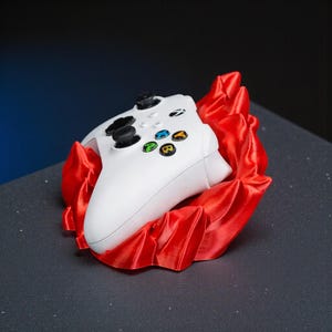 May include: A white Xbox controller rests in a vibrant red, ruffled, decorative holder. The controller features black joysticks and colored buttons. The holder is made of a shiny, textured material, creating a striking contrast.