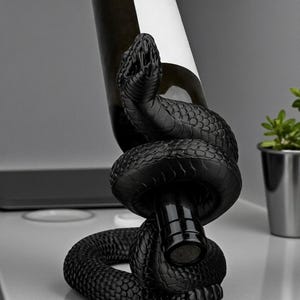 May include: A black snake wine bottle holder. The snake is coiled around a wine bottle, with the head of the snake at the top. The bottle has a white label. The holder is on a white surface.