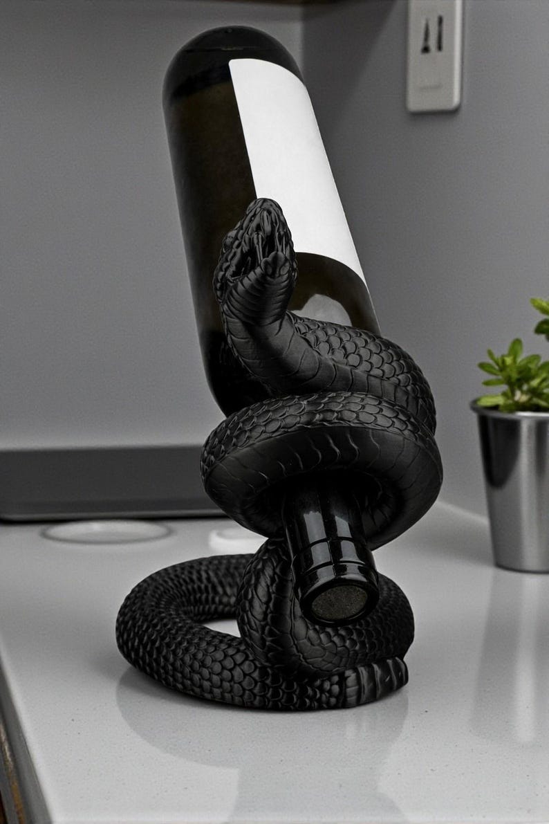 May include: A black snake wine bottle holder. The snake is coiled around a wine bottle, with the head of the snake at the top. The bottle has a white label. The holder is on a white surface.