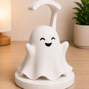 May include: A white ghost-shaped night light with a curved handle. The ghost has a smiling face and outstretched arms, sitting on a round base. The lamp is on a wooden surface, with a lamp and plant in the background.