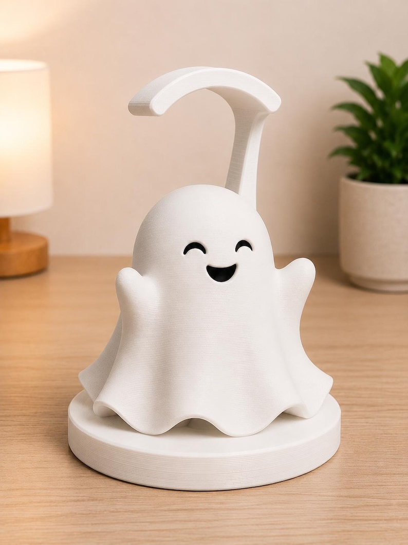 May include: A white ghost-shaped night light with a curved handle. The ghost has a smiling face and outstretched arms, sitting on a round base. The lamp is on a wooden surface, with a lamp and plant in the background.
