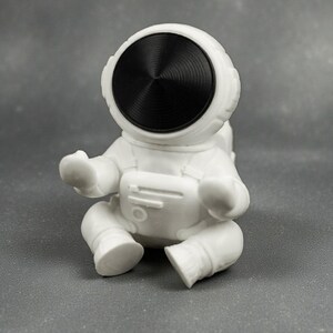 May include: A white astronaut figurine in a seated position. The astronaut has a black visor and is wearing a white spacesuit. The figurine is on a gray surface.