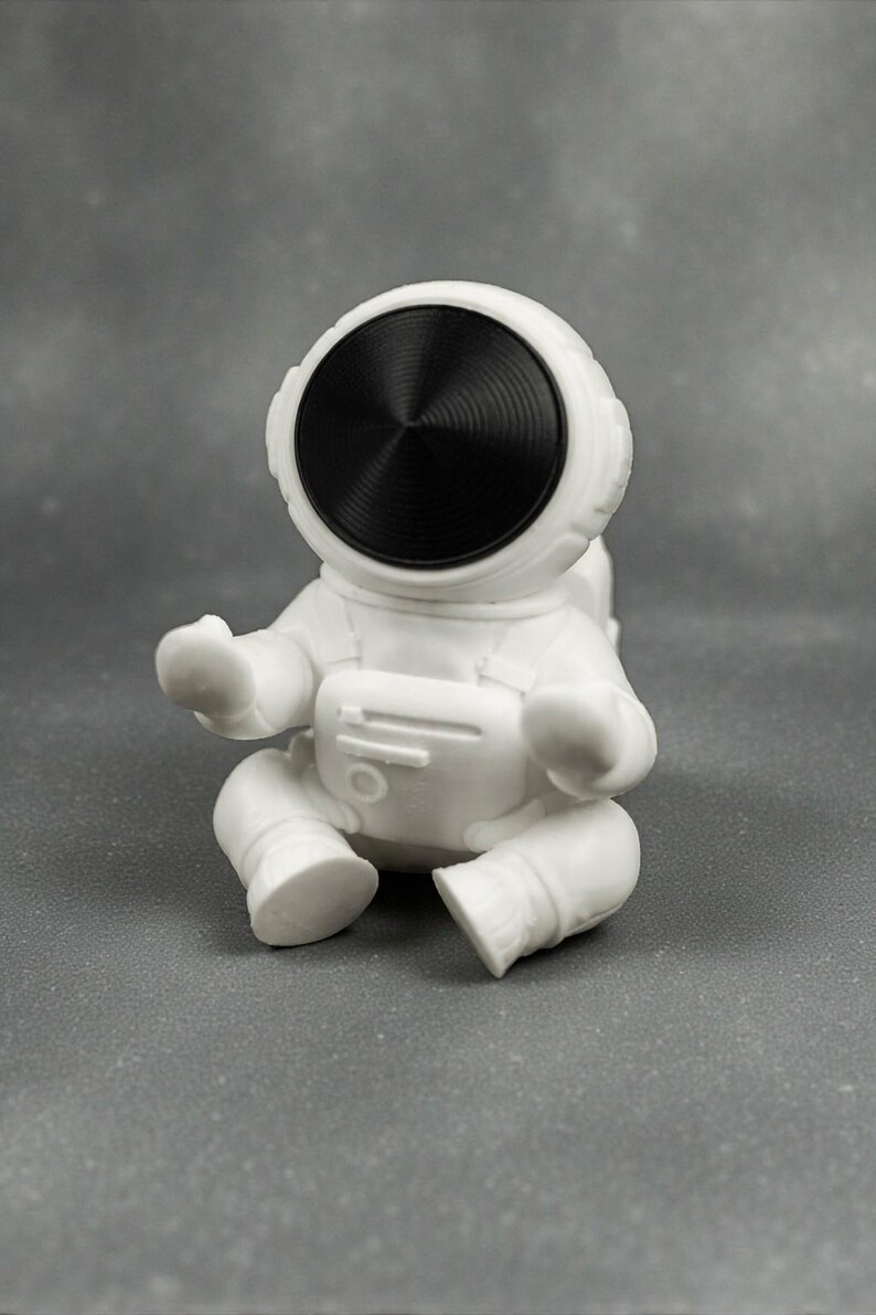 May include: A white astronaut figurine in a seated position. The astronaut has a black visor and is wearing a white spacesuit. The figurine is on a gray surface.