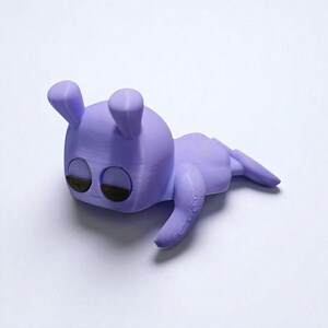 Pu&ograve; includere: Statuetta lavanda stampata in 3D di una creatura stilizzata con grandi occhi, due orecchie dritte e un corpo arrotondato. La creatura &egrave; sdraiata, con le braccia e le gambe distese. Gli occhi sono marrone scuro.