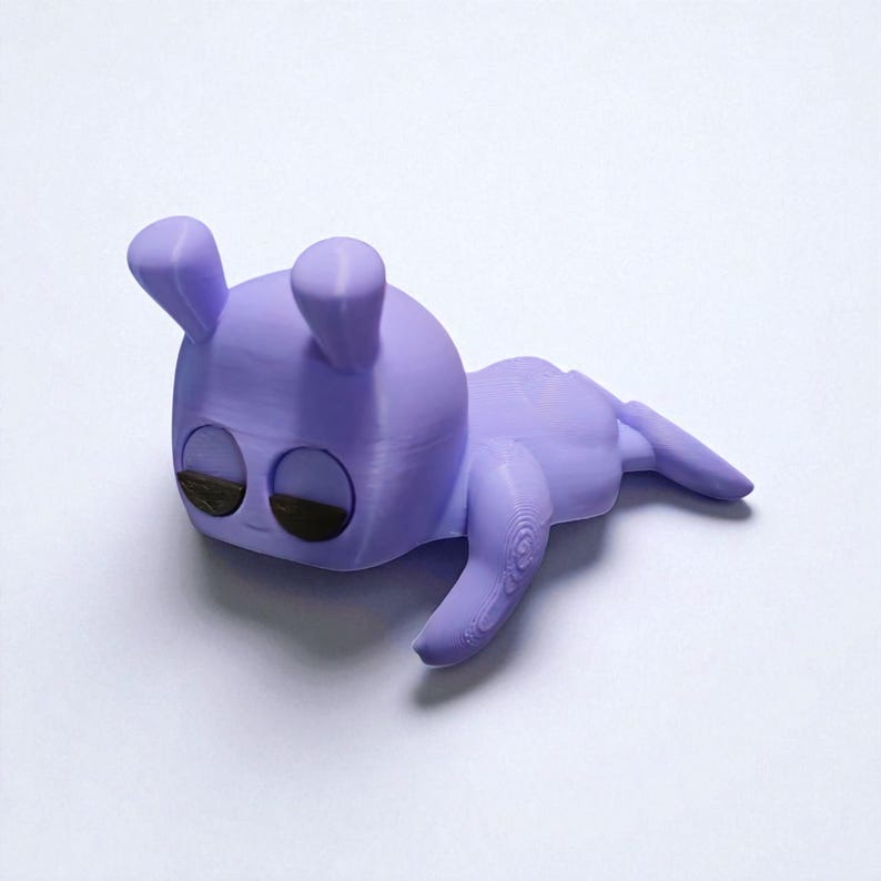 Pu&ograve; includere: Statuetta lavanda stampata in 3D di una creatura stilizzata con grandi occhi, due orecchie dritte e un corpo arrotondato. La creatura &egrave; sdraiata, con le braccia e le gambe distese. Gli occhi sono marrone scuro.