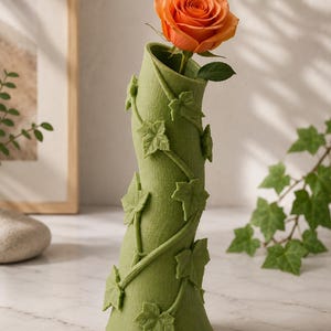 May include: A green vase with a textured surface and ivy leaf accents, holding a single orange rose. The vase has a unique, curved shape and is a decorative item for floral arrangements. The rose is in full bloom, adding a pop of color.