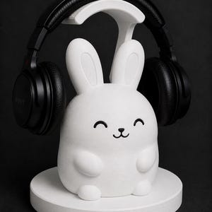 May include: A white bunny-shaped headphone stand with a smiling face and black accents. Black headphones rest on the stand. The stand is on a white circular base, all against a dark background.
